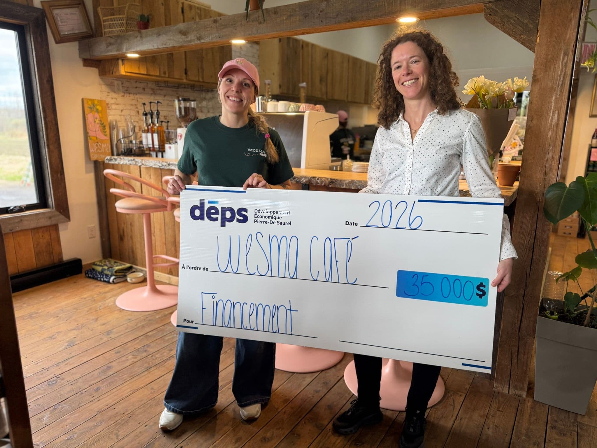 Deux femmes souriantes se tiennent à l’intérieur du Wesma Café à Sainte-Anne-de-Sorel en tenant un grand chèque symbolique du DÉPS de 35 000 $ lié à un financement provenant des Fonds locaux d’investissement et des Fonds locaux de solidarité.